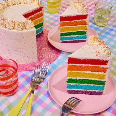 Rainbow Cake by Crumbs & Doilies with six vibrant vanilla sponge layers and smooth white sprinkle icing.